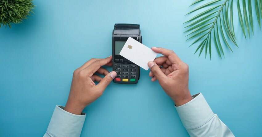 A pair of white-skinned hands hold a credit card up to a credit card machine against a teal background. Palm leaves frame the picture. environmental sustainability efforts